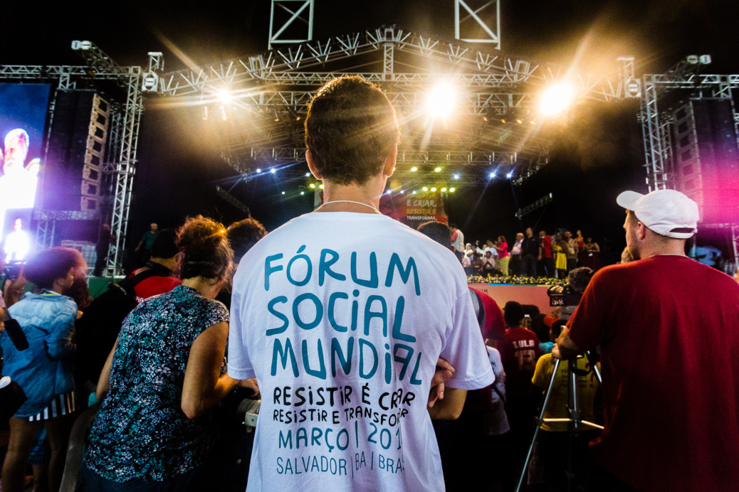 Pessoa de costas, com logo do FSM na camiseta, e ao fundo o palco do Ato Democracias, no estádio do Pituaçu