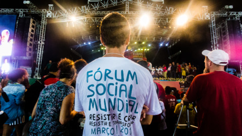 Pessoa de costas, com logo do FSM na camiseta, e ao fundo o palco do Ato Democracias, no estádio do Pituaçu