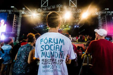 Pessoa de costas, com logo do FSM na camiseta, e ao fundo o palco do Ato Democracias, no estádio do Pituaçu